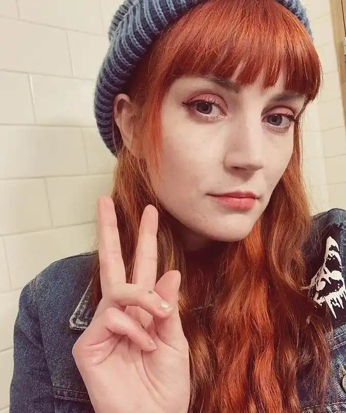 Britt is a white woman with long red hair with a fringe. She is wearing a cure beanie and giving a peace sign. She’s wearing makeup and looking lovely